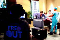 A person in a black shirt with 'FRE OUT' text watches a TV showing a medical scene in a room.