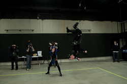 A person in a motion capture suit performs an upside-down aerial stunt in a studio with cameras and crew.