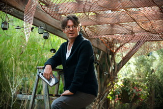 A man with glasses and a black cardigan sits on a wooden ladder under a rustic mesh canopy.