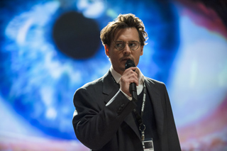 A man with glasses and a mustache speaks into a microphone against a large blue eye backdrop.