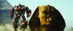 A large, colorful mechanical robot stands in a desert landscape next to the Great Sphinx.