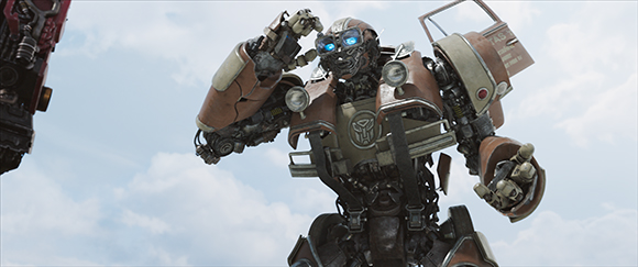 A large tan and brown mechanical robot with glowing blue eyes stands against a cloudy sky.