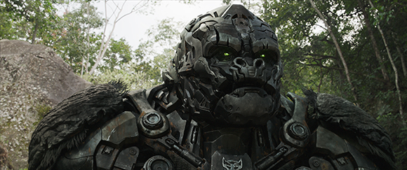A close-up of a large, dark metallic robotic creature with glowing green eyes in a forest.