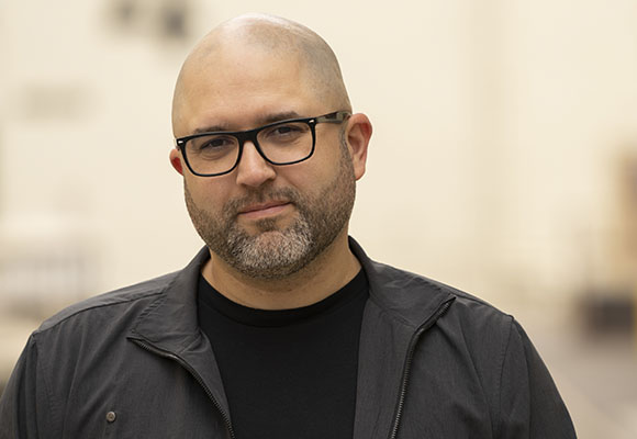 A bald man with glasses and a salt-and-pepper beard wearing a black shirt and jacket.