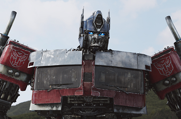 A large blue and red robot with glowing blue eyes stands against a light sky.