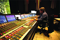 A man sits at a large, illuminated professional audio mixing console in a dark studio.