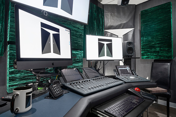 A studio desk with dual monitors, a large mixing console, studio monitors, and acoustic panels.