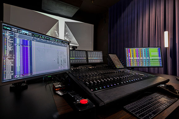 A large audio mixing console sits before several computer monitors displaying digital audio workstation software.
