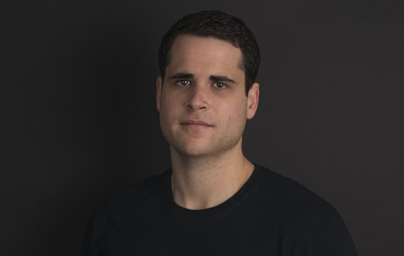 A man with short dark hair and a black t-shirt looking forward against a dark background.