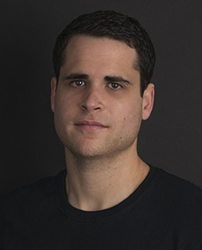 A man with short dark hair and a black t-shirt looking forward against a dark background.