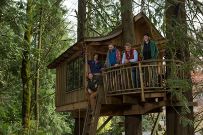 Five men posing on the wooden balcony of a multi-level treehouse built into tall evergreen trees.