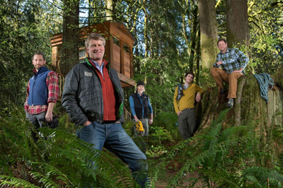 Five men pose in a lush, green forest setting near a wooden treehouse.