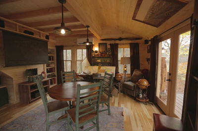 A rustic living room with wood walls, a round dining table, and glass doors leading to a deck.