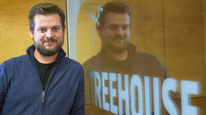 A man with a beard wearing a blue zip-up sweater standing next to a glass door reflecting his image.