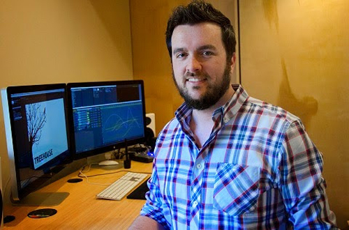 A man with a beard wearing a blue and red plaid shirt sits in an office with two computer monitors.