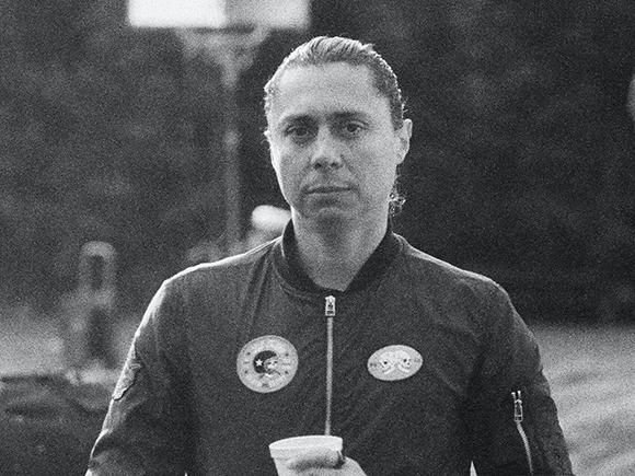 Black and white portrait of a person with hair pulled back, wearing a dark bomber jacket with circular patches.