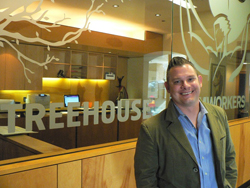 A smiling man in a green blazer and blue shirt stands in front of a glass wall with Treehouse branding.