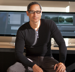 A man with glasses and a black shirt sits in front of large computer monitors displaying editing software.