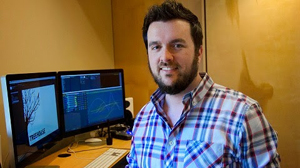A man with a beard wearing a plaid shirt smiles in an office with two computer monitors.