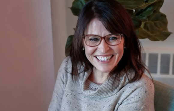A smiling woman with dark hair and glasses wearing a grey turtleneck sweater.