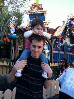 A man carries a young child on his shoulders in front of a whimsical, colorful building.