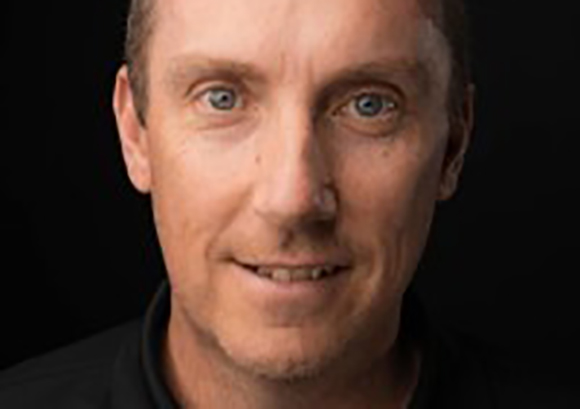 Close-up portrait of a man with light eyes and a slight smile against a dark background.