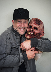 A smiling man in a black cap and grey jacket holds a large, bloody zombie mask to his face.