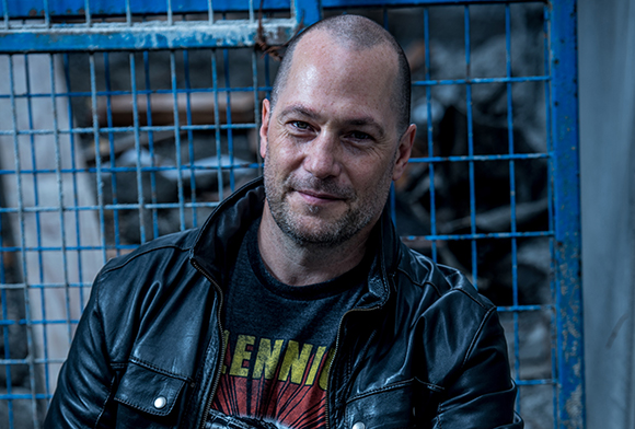 A man with short hair and stubble wearing a black leather jacket and a graphic t-shirt.