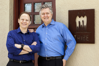 Two men smiling, one in a dark blue shirt and one in a light blue shirt, standing by a door and a sign.