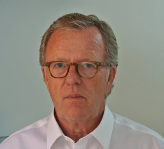 A man with short graying hair and brown-rimmed glasses wearing a white collared shirt.