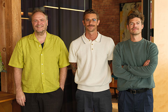 Three men standing in a row: one in a lime shirt, one in a white polo, and one in a teal sweatshirt.