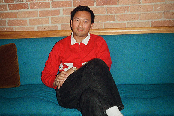 A man wearing a red sweater and black pants sits on a teal couch against a brick wall.