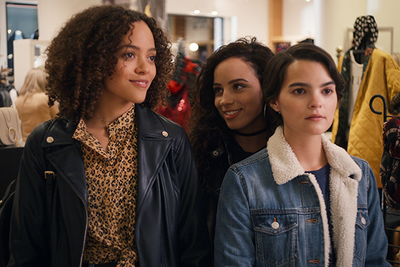 Three young women looking forward while standing in a brightly lit clothing boutique.