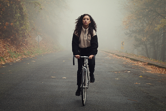 A woman in a black and tan jacket rides a bicycle toward the camera on a foggy, tree-lined road.