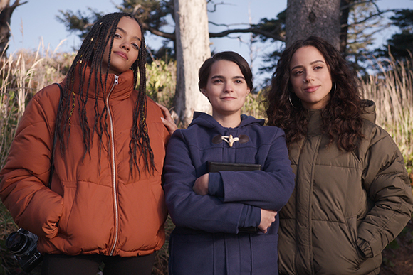 Three young women posing together outdoors wearing puffer jackets and a blue coat.
