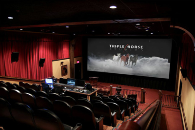 Interior of a dark movie theater with red curtains and a large screen showing horses running through clouds.