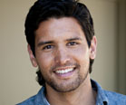 A smiling man with dark hair and light stubble wearing a blue shirt.