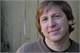 A man with light brown hair smiling in front of a tan wooden wall.