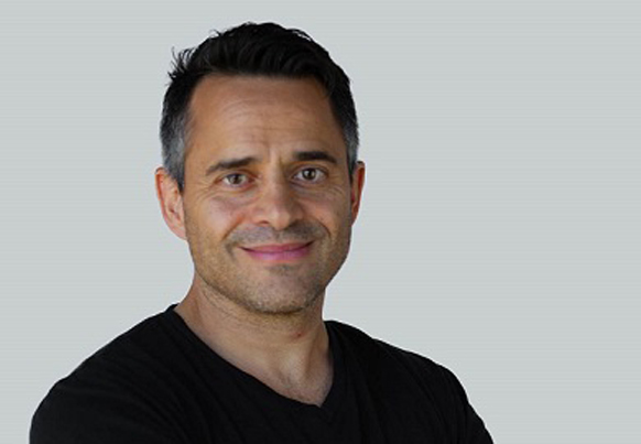 A man with short dark hair and light stubble smiling against a plain gray background.