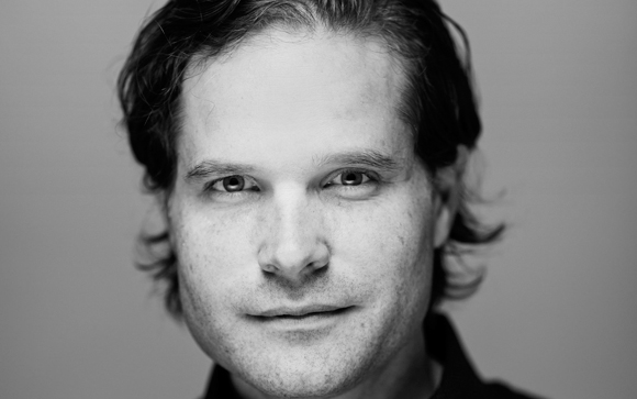 Black and white close-up portrait of a man with wavy hair looking directly at the camera.