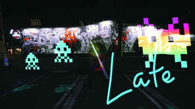 A person stands before a large black and white mural with colorful pixelated ghost overlays and the word Late.