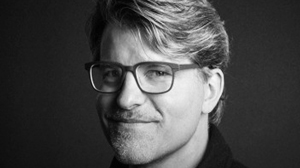 Black and white headshot of a man with glasses and a slight smile against a dark background.