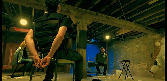 A person sits in a chair with hands tied behind their back, facing another seated person in a dark, rustic room.