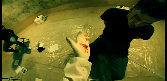 A person in a dark shirt leans over a plastic-covered floor near a hand in a blood-stained white glove.