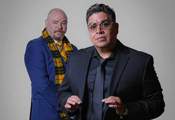 Two men in formal attire; one in a black suit and tie, the other in a blue suit and yellow plaid scarf.