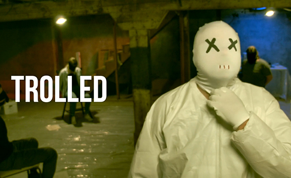 A person in a white mask with X eyes and a white suit stands in a dark room with 'TROLLED' text overlaid.