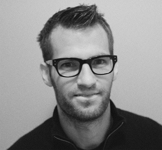 Close-up headshot of a man with short hair, wearing thick-rimmed glasses and a dark collared shirt.