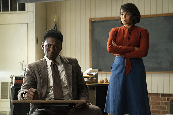 A man in a brown suit sits at a desk looking serious while a woman in a red shirt stands behind him.