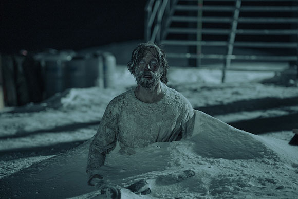 A man with a disheveled appearance sits partially submerged in a large pile of snow at night.