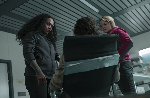 Two women stand over a seated person in a dimly lit room.